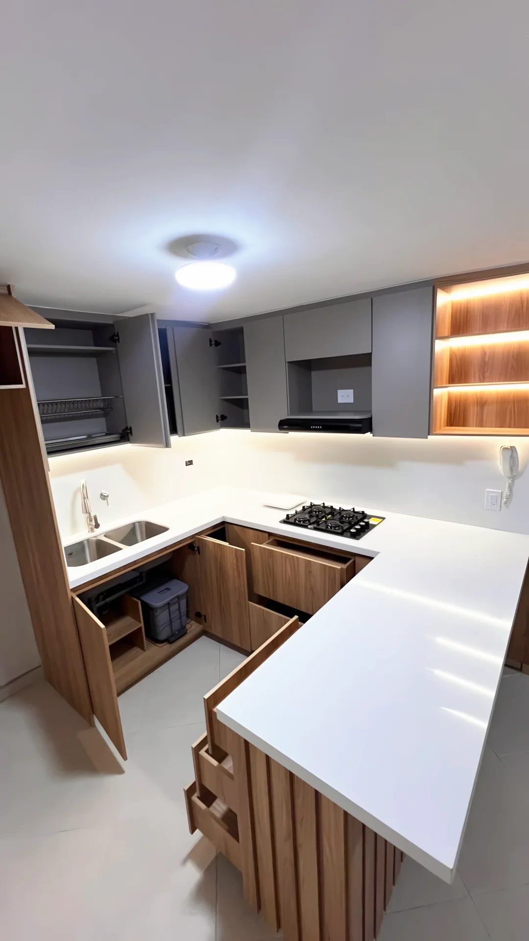 Cocina en U terminada: muebles en madera tono nogal, alacenas grises, repisas con luz cálida, mesón blanco y barra en listones, con cajones abiertos